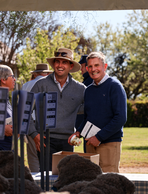 Two men smiling at an outdoor wool or livestock event, standing behind a table with wool samples and labelled stands.