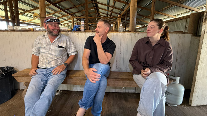 Woolgrower Michael Edmonds sits with Deakin University researchers Dr. Andrew Oxley and Madison Bone on a wooden bench inside a rustic shearing shed during a flystrike research visit at Haylands, Armidale. (https://www.wool.com)