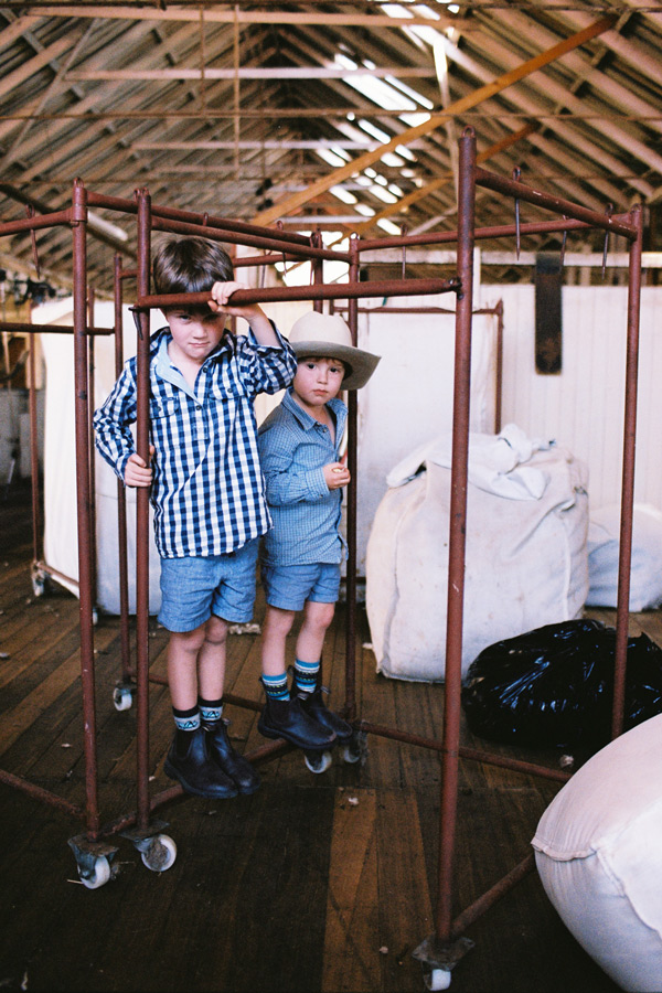 Two young boys in gumboots standing in a sheep shearing shed with wool bales and equipment