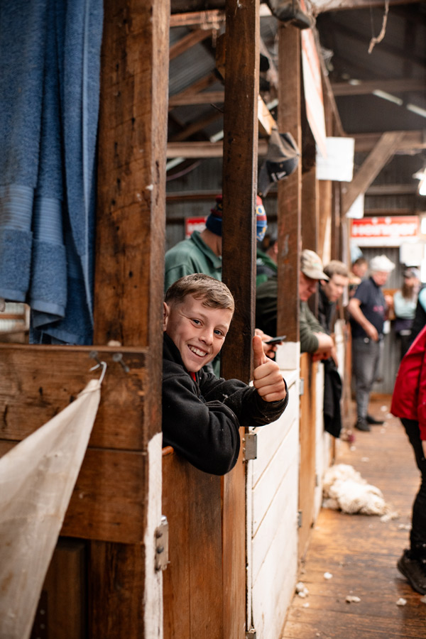 Young girl carrying a large fleece with others working in a shearing shed in the background