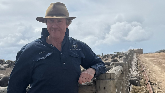 Simon Fowler of Chilwell Farms, Western Australia, wearing a wide-brimmed hat and navy work shirt, leaning against a fence with a large mob of Merino sheep in a containment feeding area behind him.